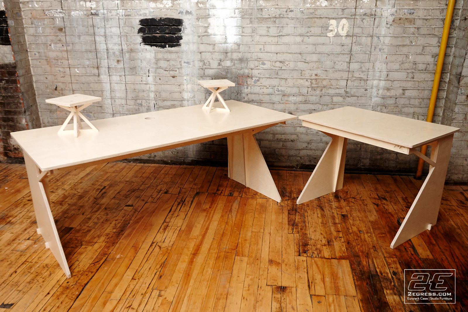 Facet desk | pair of matching desks with X-shaped embedded monitor stands, front view against a brick wall | 2Egress